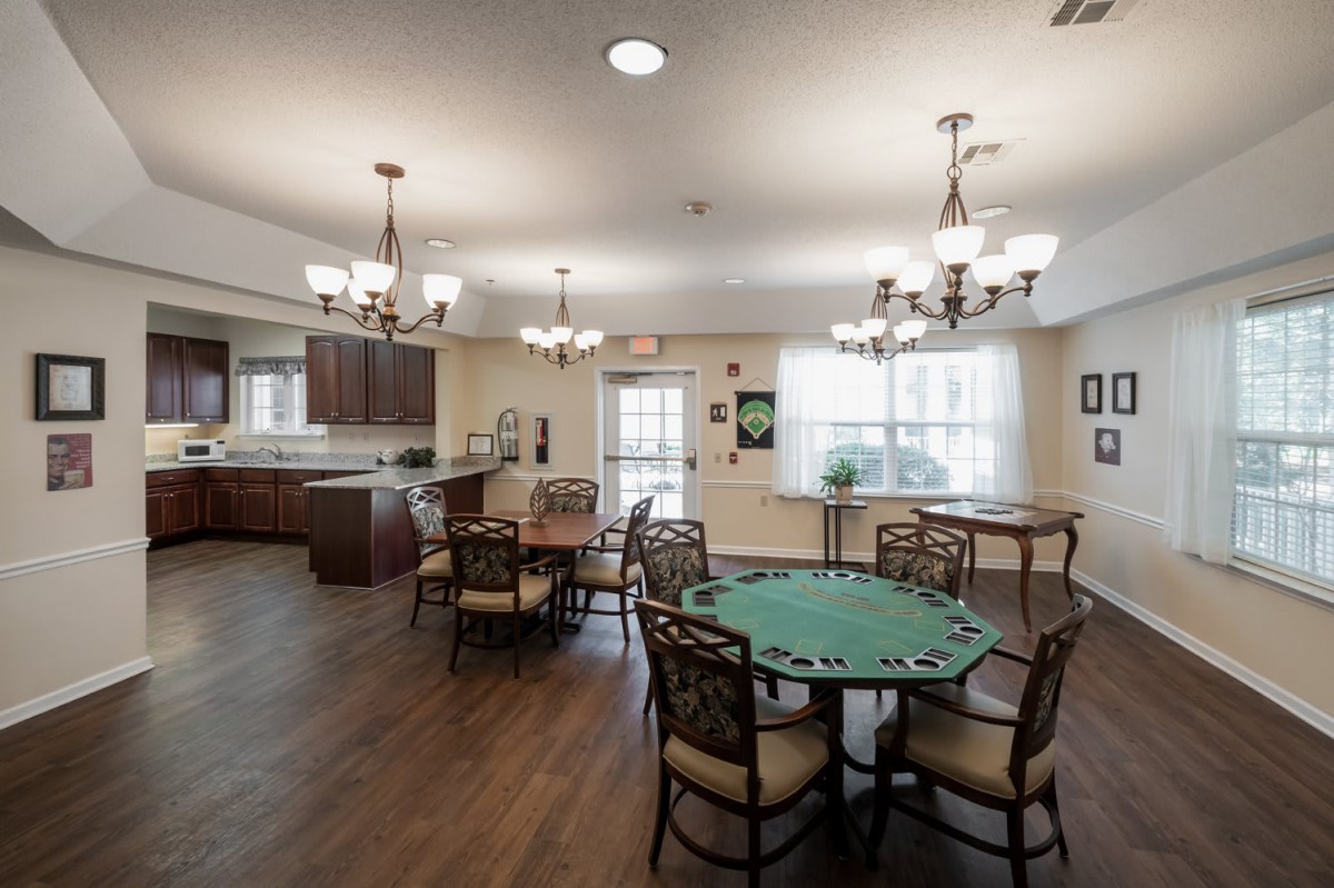 A cozy dining area with a poker table and a view into the kitchen, featuring elegant chandeliers and hardwood flooring.