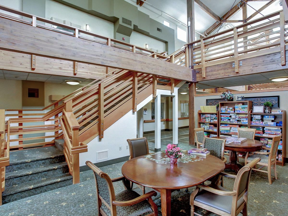 A cozy library interior featuring a round table with chairs, wooden bookshelves, and a staircase leading to an upper level.