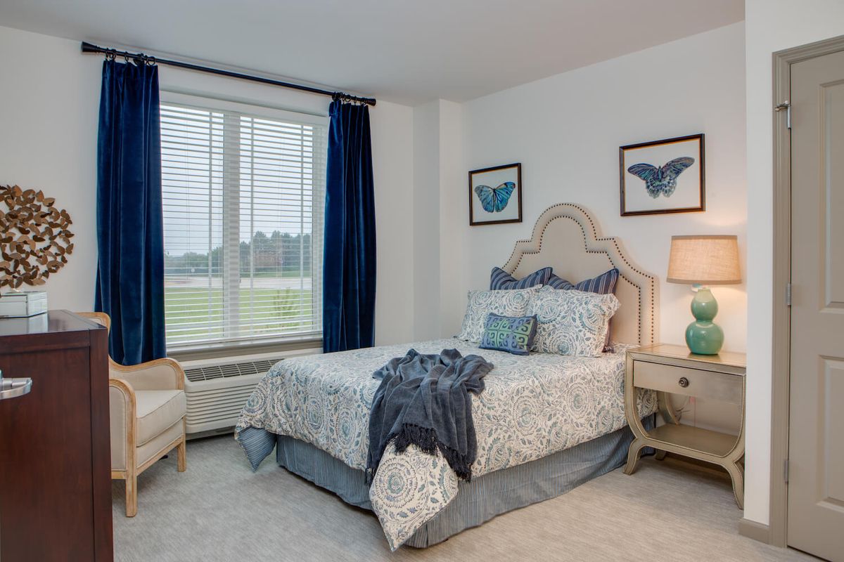 A cozy bedroom featuring a neatly made bed with patterned bedding, flanked by side tables and lamps, complemented by blue curtains and decorative wall art.