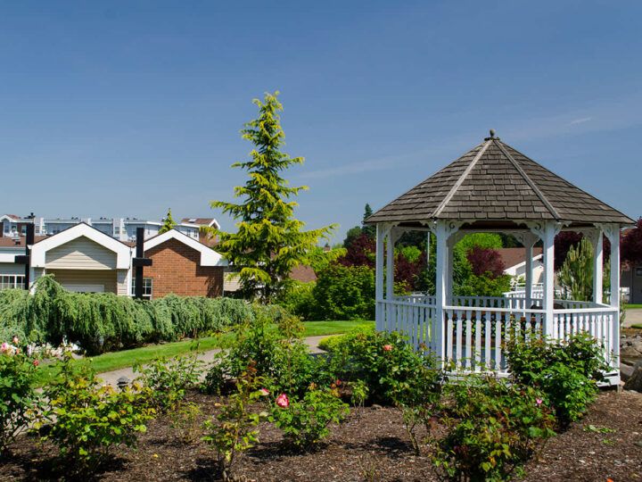 A picturesque garden with a white gazebo and houses in the background under a clear blue sky.