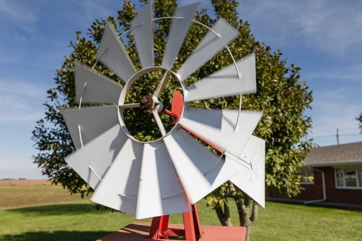 A metal windmill stands outdoors, surrounded by trees and grass.