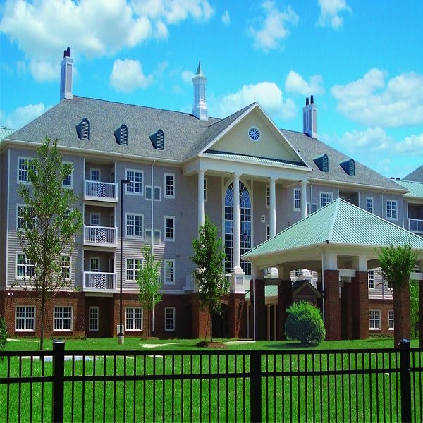 A view of a multi-story residential apartment building with balconies, surrounded by green lawns and bordered by a black fence under a partially cloudy sky.
