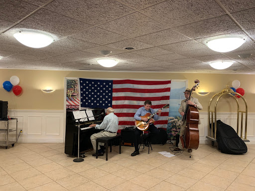 A lively music performance featuring a pianist, guitarist, and bassist in front of an American flag backdrop.