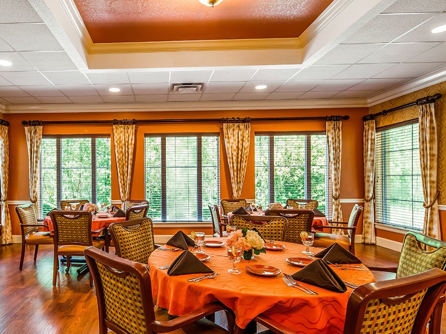 A warmly decorated dining room with round tables set for a meal, featuring large windows with views of greenery.