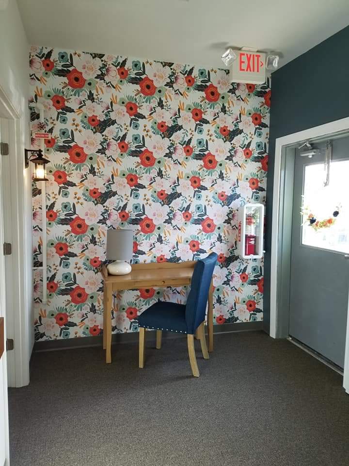 A cozy corner with vibrant floral wallpaper, featuring a small wooden desk and a blue chair, complemented by a stylish lamp.
