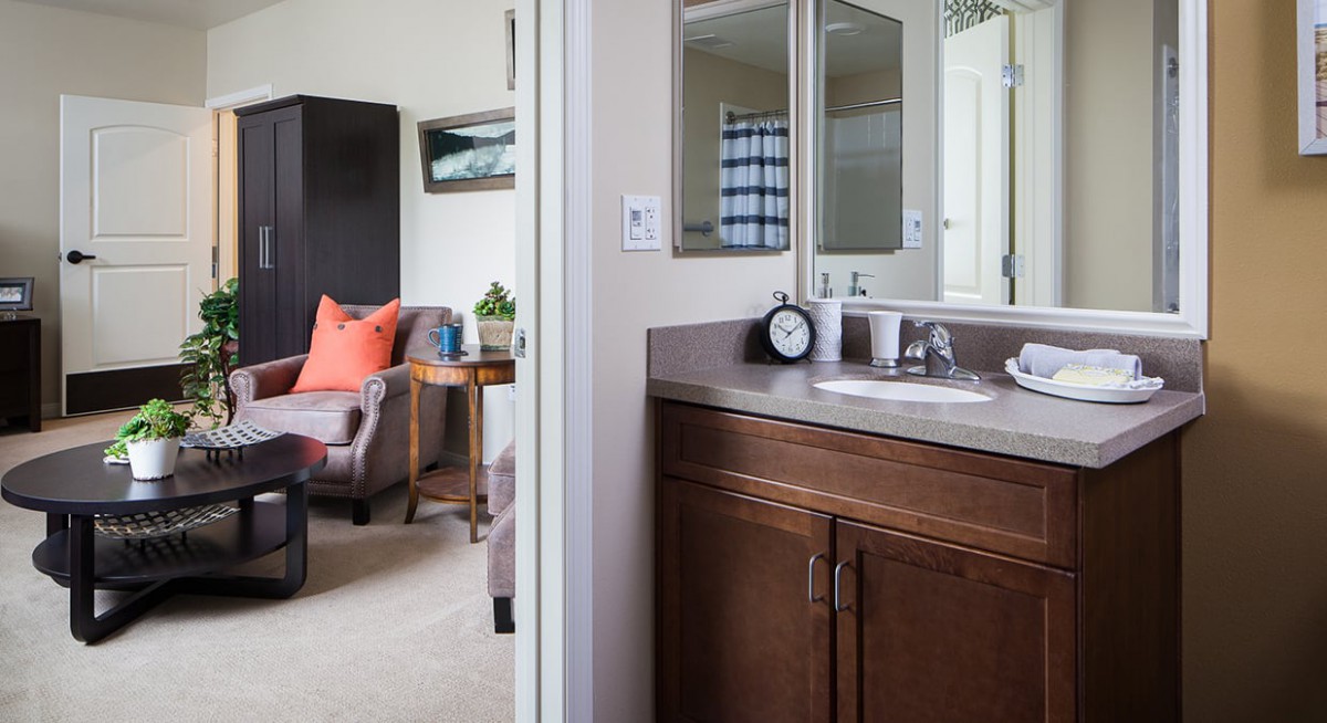 A cozy bedroom with an adjoining bathroom featuring a sink and mirror, along with a comfortable chair and side table.