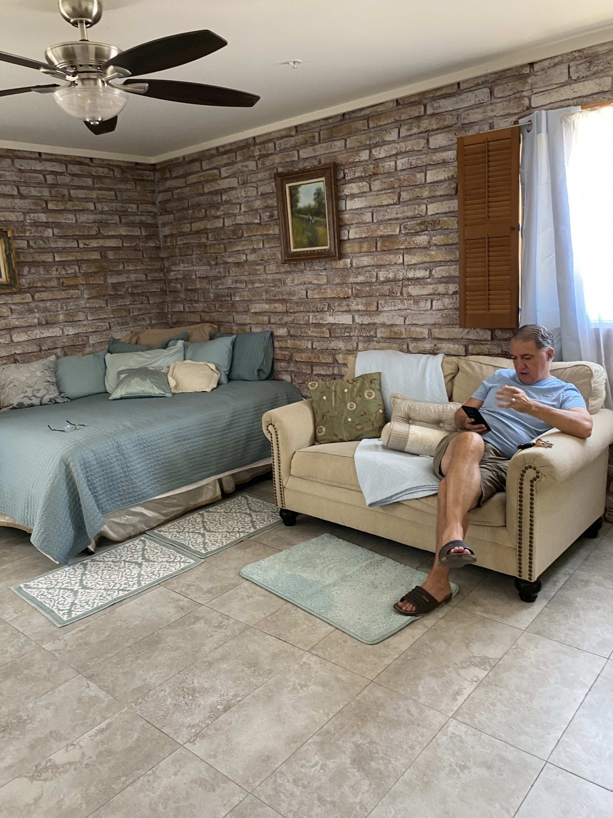 A cozy living room with a man reading on the sofa, adjacent to a bed, featuring brick walls and ample natural light.