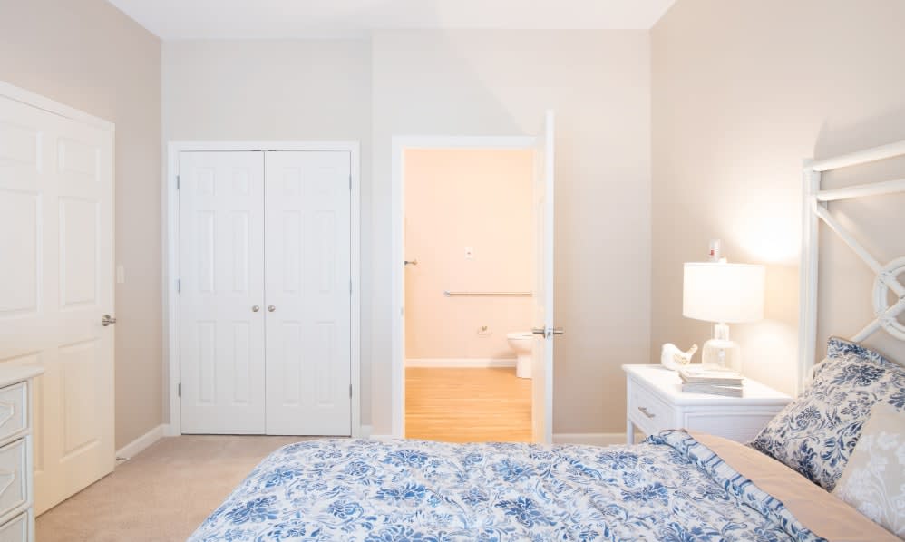 A cozy bedroom featuring a neatly made bed with blue patterned bedding, a bedside lamp on a nightstand, a closet with double doors, and an adjoining bathroom.
