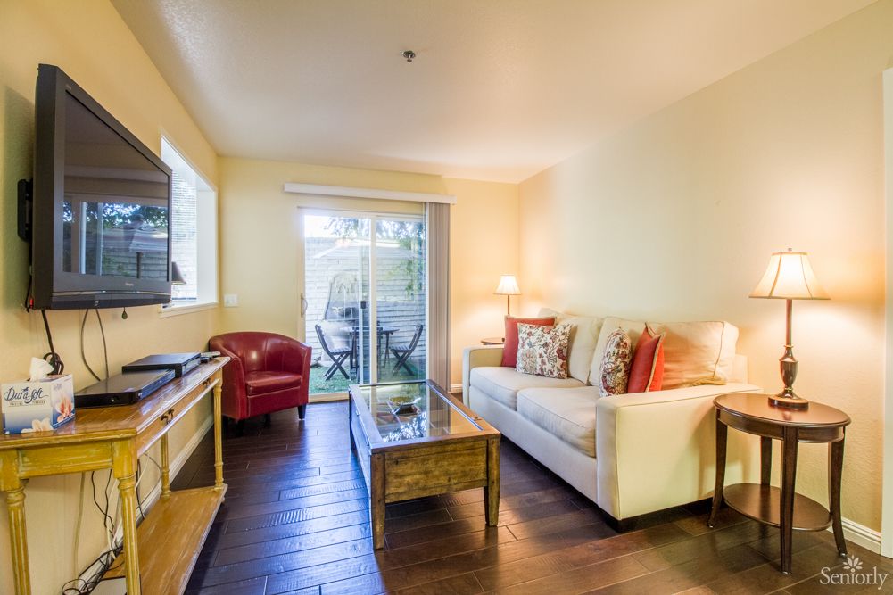 A cozy living room with a sofa, armchair, and coffee table facing a television. A sliding door opens to a small patio, enhancing the warm ambiance.