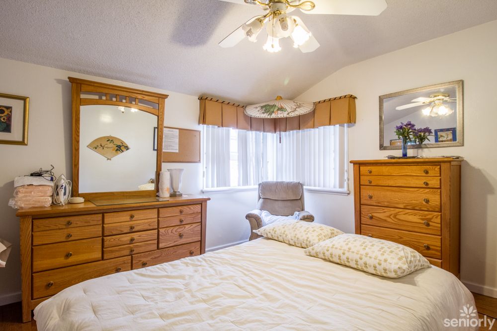 A cozy bedroom featuring wooden furniture, including a dresser with a mirror and a bed. The room is well-lit with natural light from the windows and has a ceiling fan for ventilation.