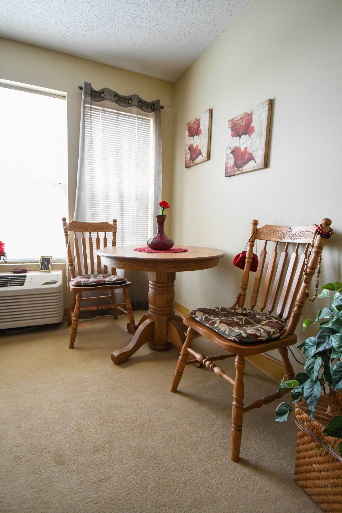 A cozy dining nook with a round wooden table surrounded by two matching chairs, adorned with floral wall art and a soft curtain filtering natural light.