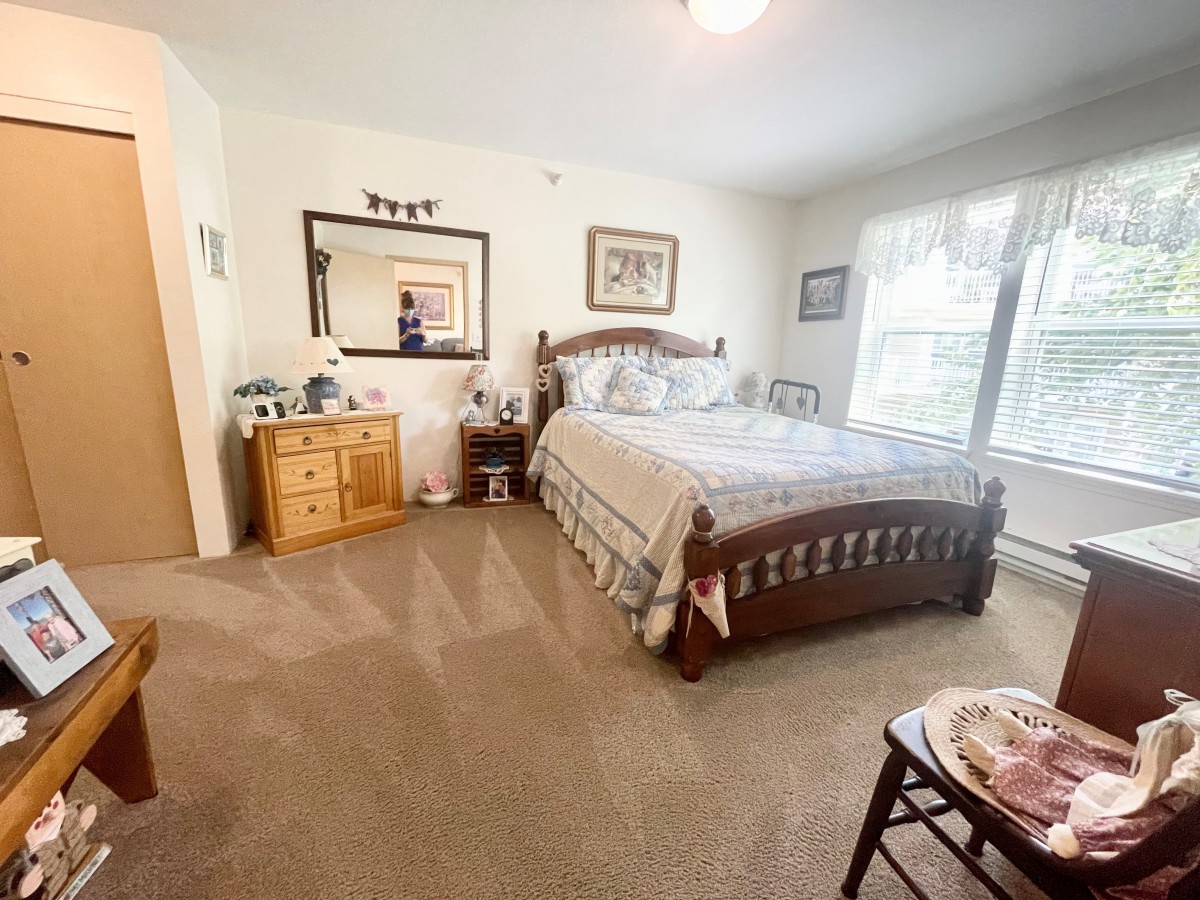 A cozy bedroom with a wooden bed frame, dresser, and large mirror, featuring soft carpeting and bright windows.