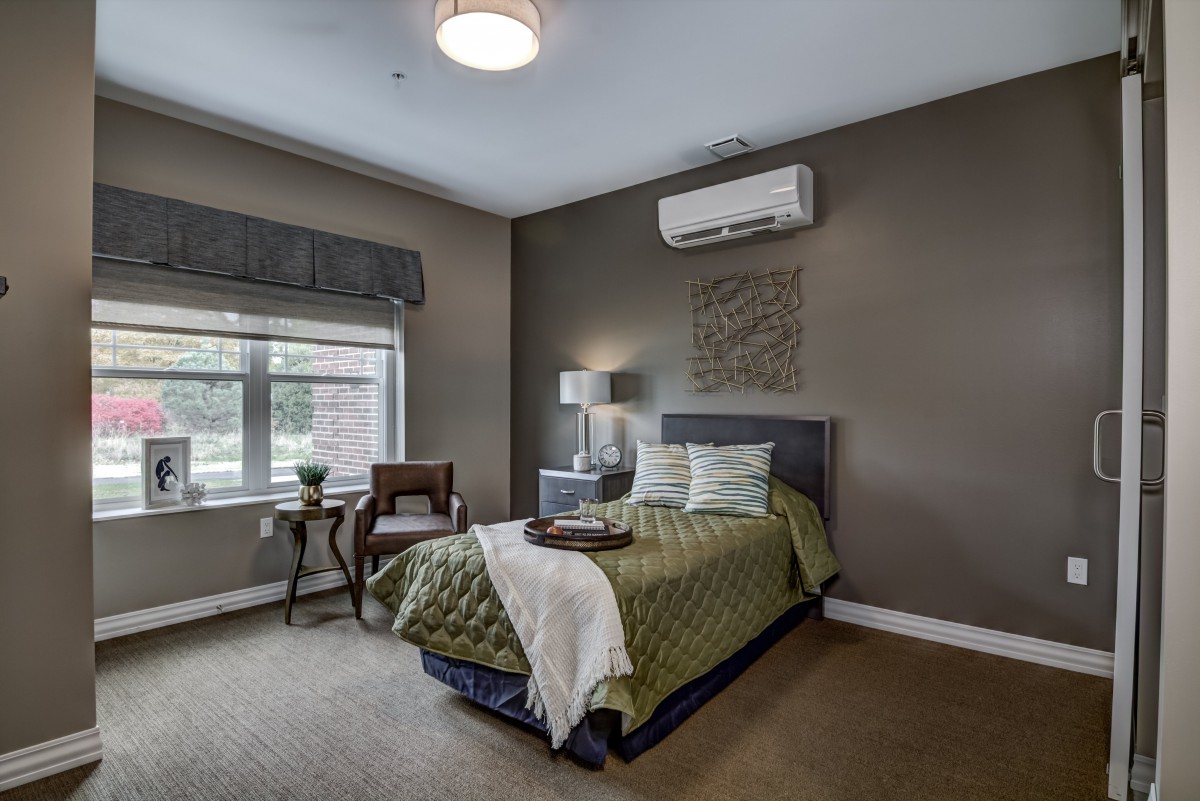 A modern bedroom with a single bed featuring green bedding, an air conditioner above the headboard, and a window allowing natural light in.
