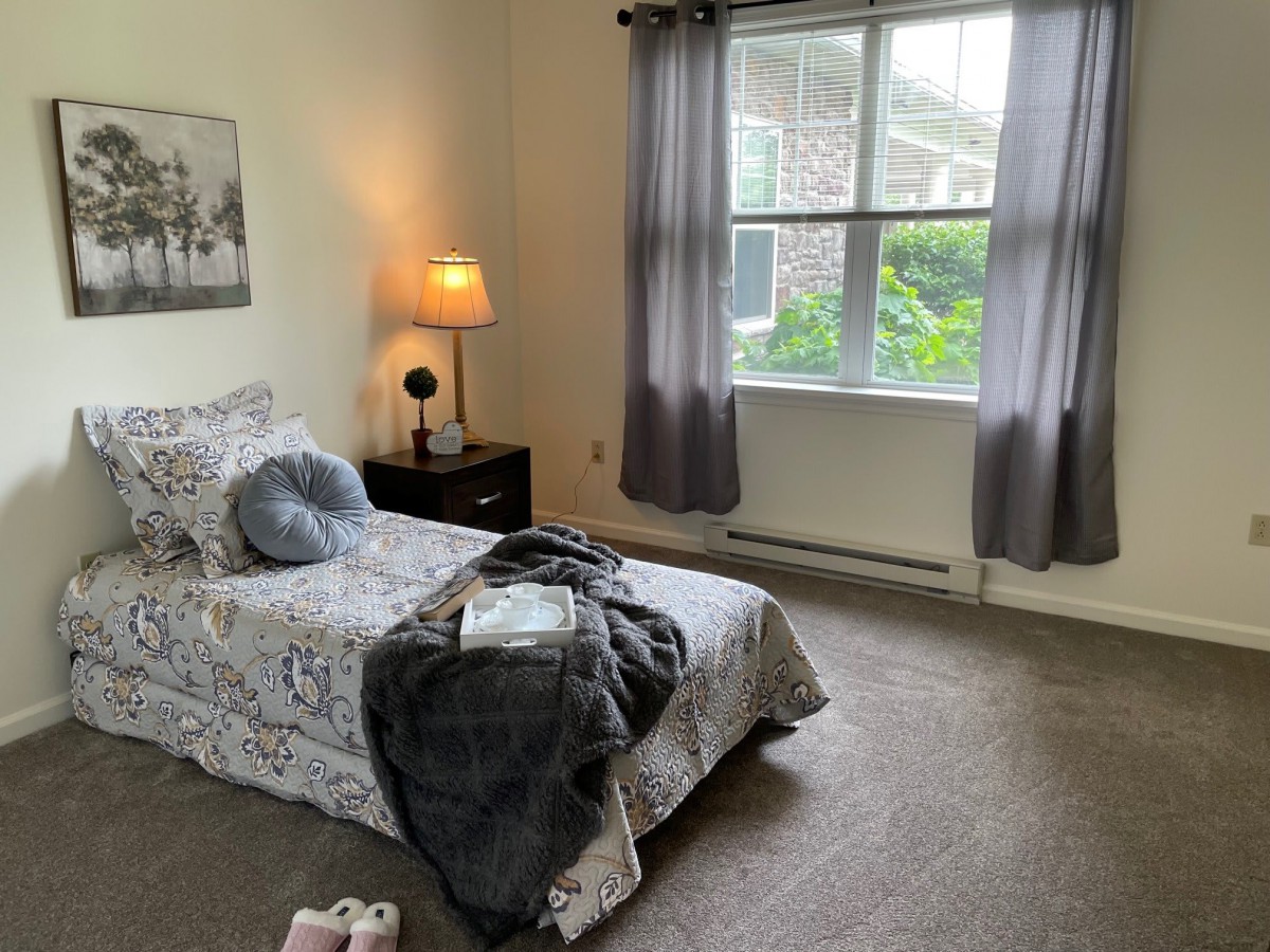 A cozy bedroom with a single bed, bedside lamp, and a large window letting in natural light.