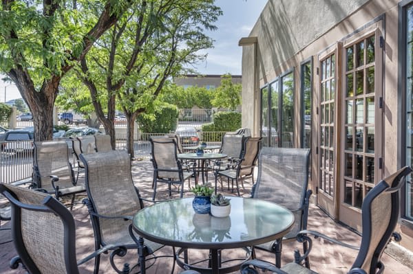A sunny outdoor patio with tables and chairs surrounded by leafy trees.