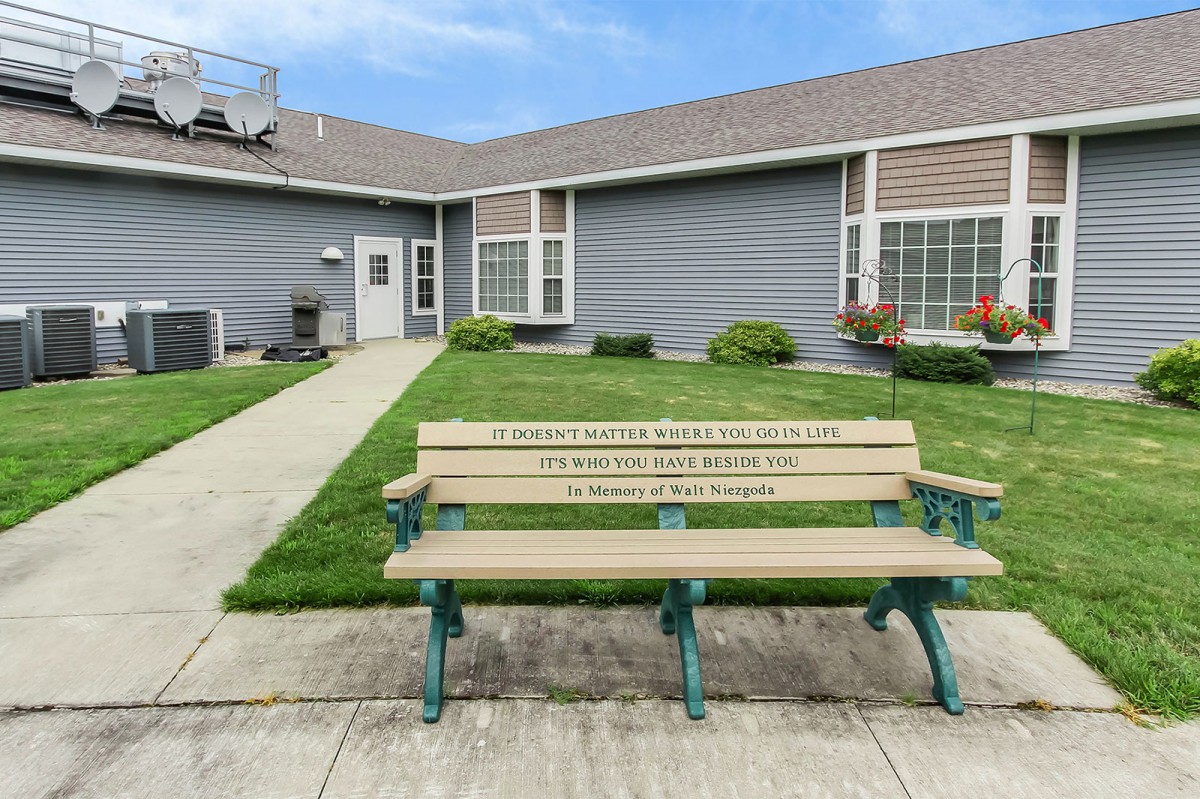 A memorial bench with an engraved plaque placed in front of a building and green lawn.