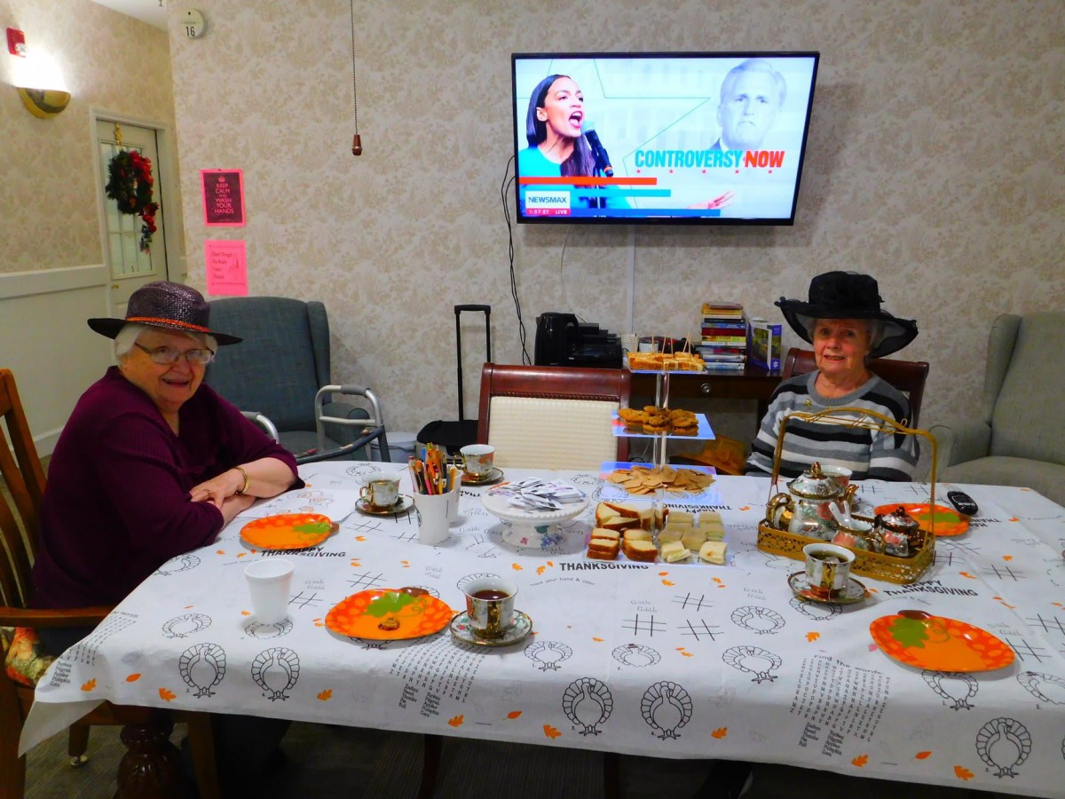 Two people enjoying afternoon tea at a table with snacks and hats, while a television is playing in the background.