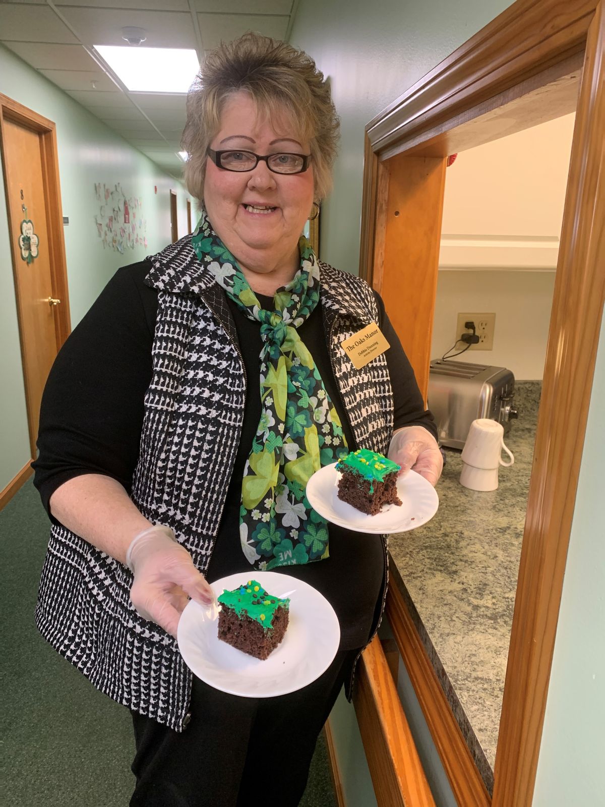 A smiling person holding two plates of cake with green frosting indoors.