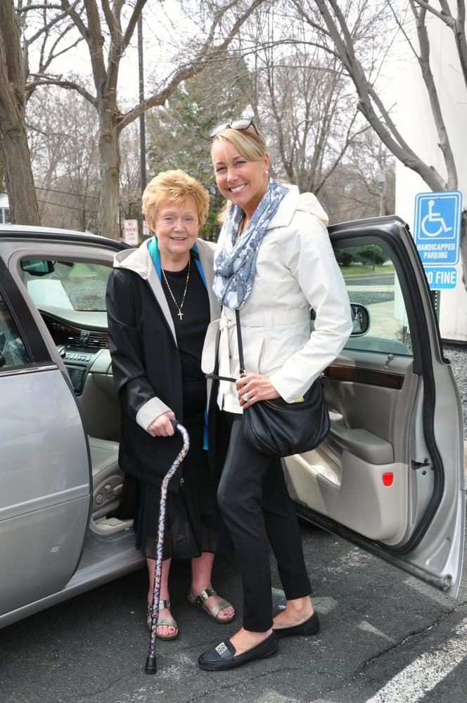 An elderly woman with a cane stands beside a younger woman near a car, both smiling.