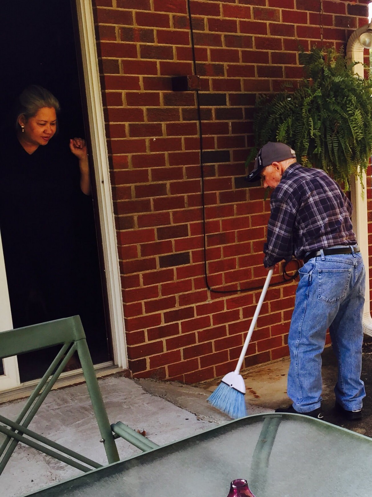 An elderly man is sweeping the porch while a woman watches from the doorway.