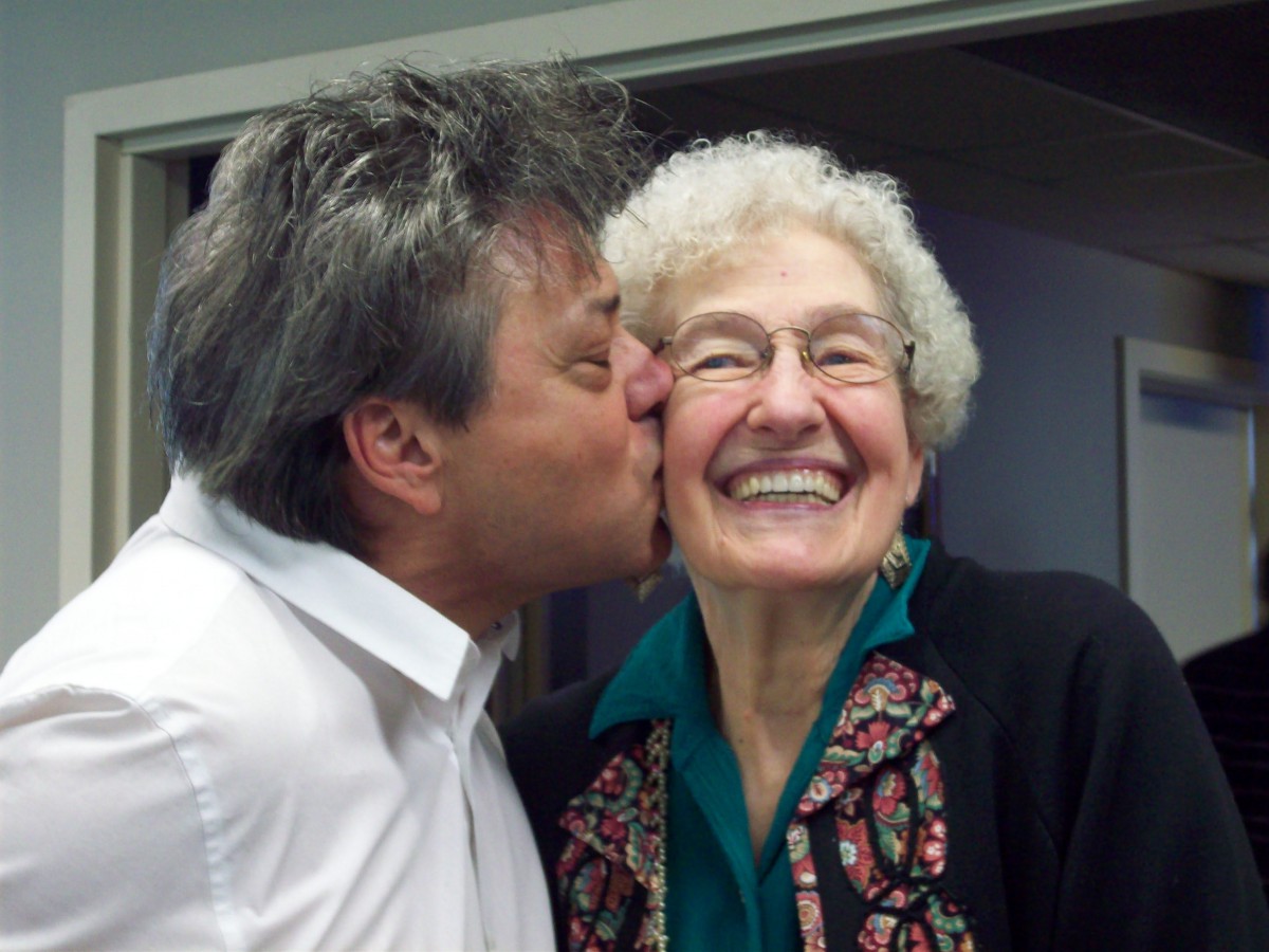 A man kisses an elderly woman's cheek, both smiling warmly.