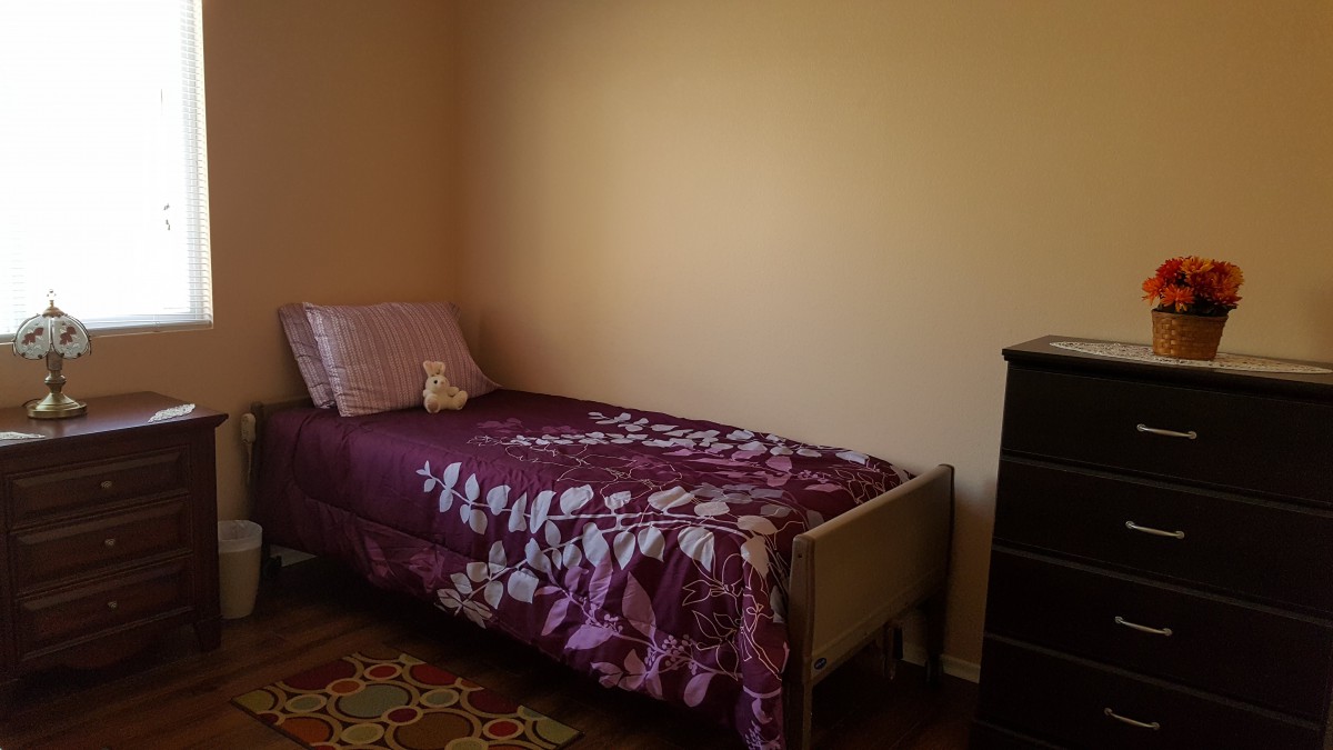 A minimalist bedroom with a single bed adorned with purple floral bedding and a pillow, next to a dark-colored dresser.