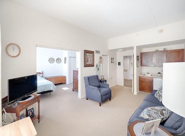 A cozy living space with a view into the bedroom, featuring a sofa, armchair, and kitchenette.