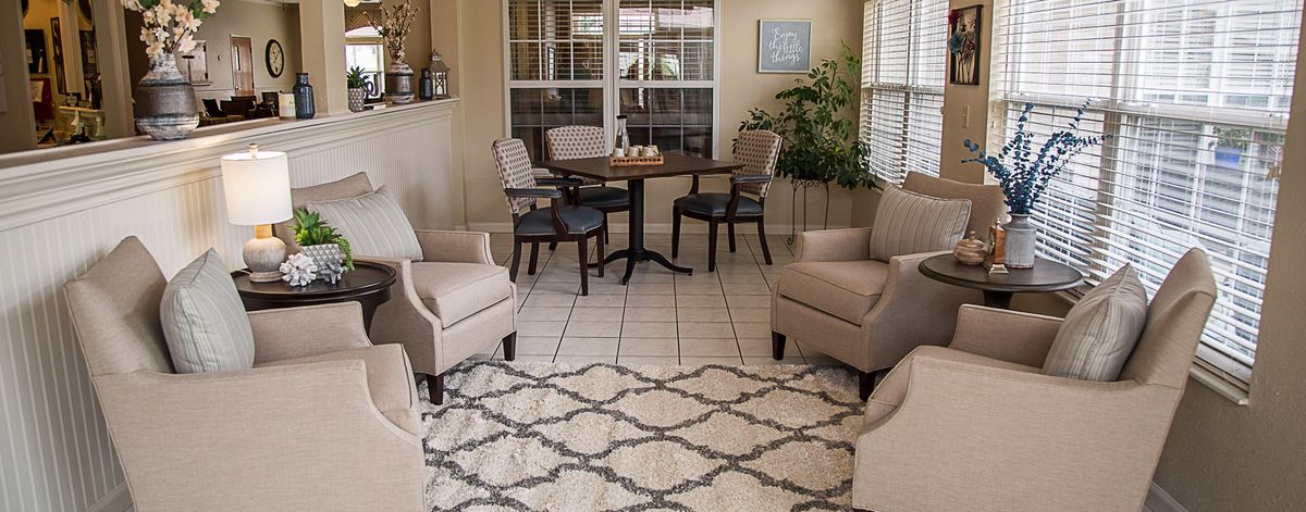 A cozy living room with a seating area, including upholstered chairs and a round table, complemented by a patterned rug and indoor plants.