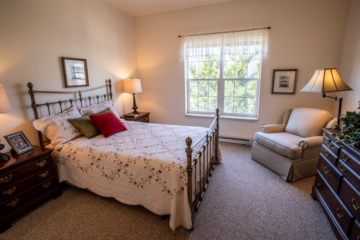 A cozy bedroom with a decorative bed, a window letting in natural light, and a comfortable chair.