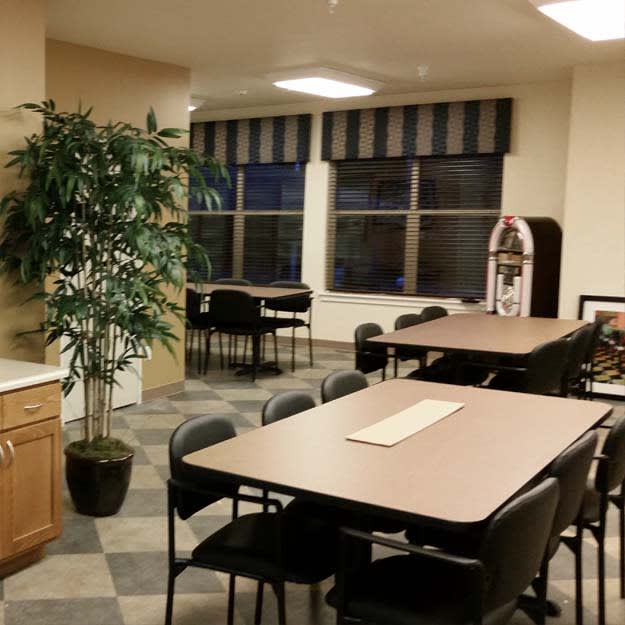 A cozy dining area with multiple tables and chairs, featuring a kitchenette and plants near the windows.