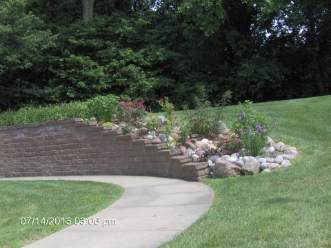 A landscaped garden featuring a stone pathway, brick retaining wall, and various plants.