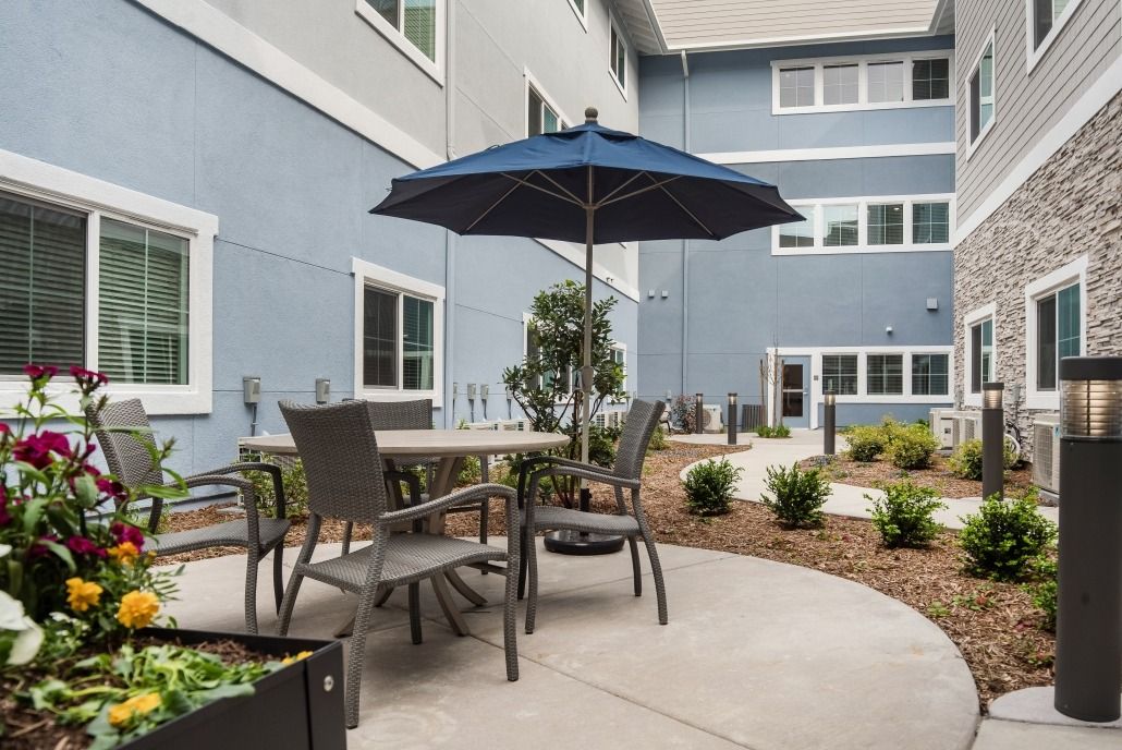 A patio area with a table, chairs, and an umbrella set against a landscaped garden backdrop.
