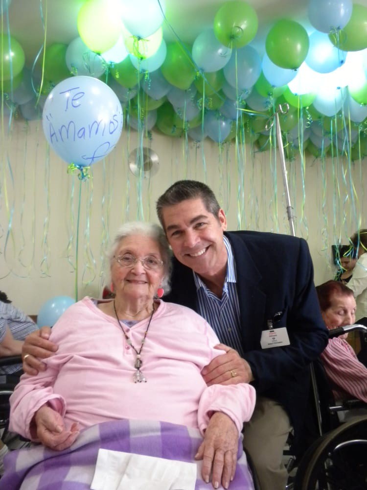 An elderly woman and a man smiling at a celebration with balloons in the background.