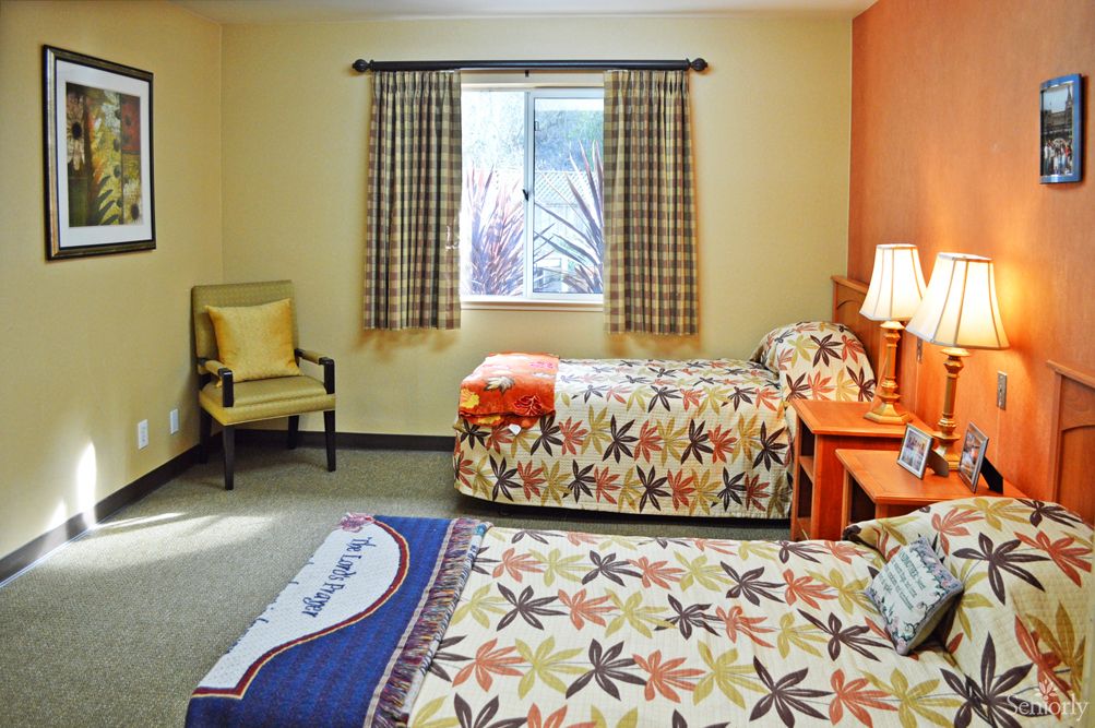 A cozy bedroom with two twin beds featuring colorful floral bedspreads, a window with checkered curtains, a lamp on the bedside table, and an upholstered chair in the corner.