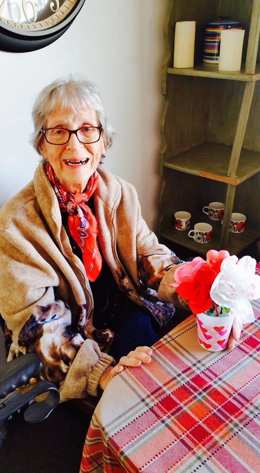 An elderly woman with glasses and a scarf sits at a table, smiling warmly with a bouquet of flowers in front of her.