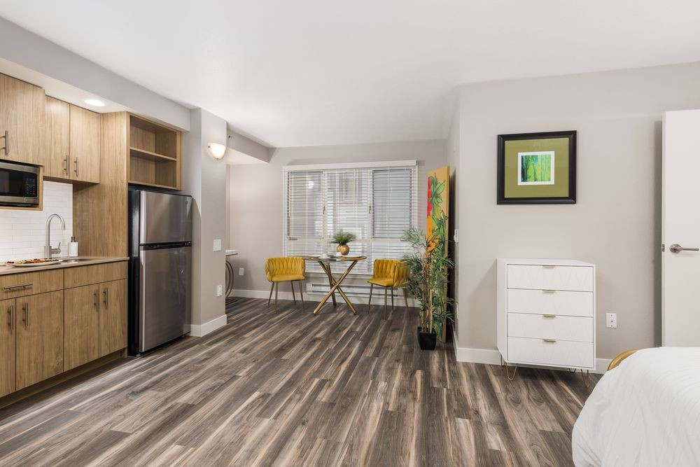 A modern apartment interior featuring a cozy dining area with yellow chairs, a stainless steel fridge, wood flooring, and an indoor plant.