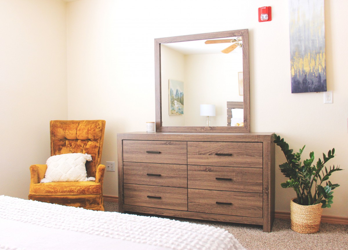 A cozy bedroom featuring a wooden dresser with a mirror, alongside an upholstered chair and a potted plant.