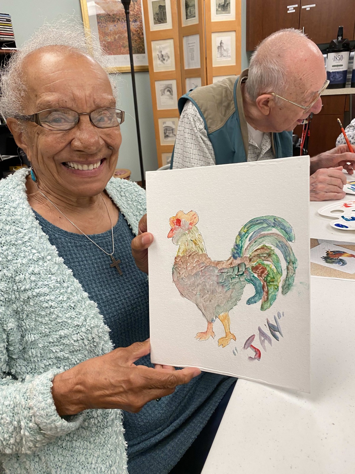 An elderly woman smiles while showing her painting of a rooster in an art class.