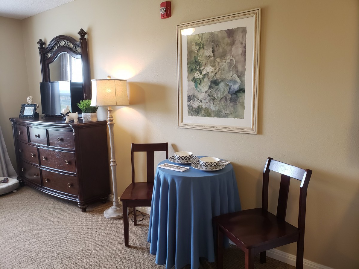 A cozy interior space featuring a small table with a blue tablecloth, two wooden chairs, a floor lamp, and framed artwork on the wall.