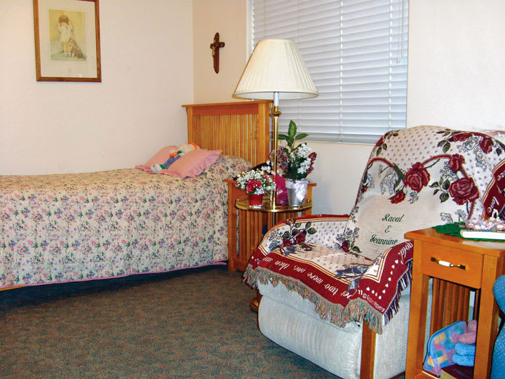 A cozy bedroom featuring a neatly made bed, an armchair with a decorative blanket, and a table lamp.
