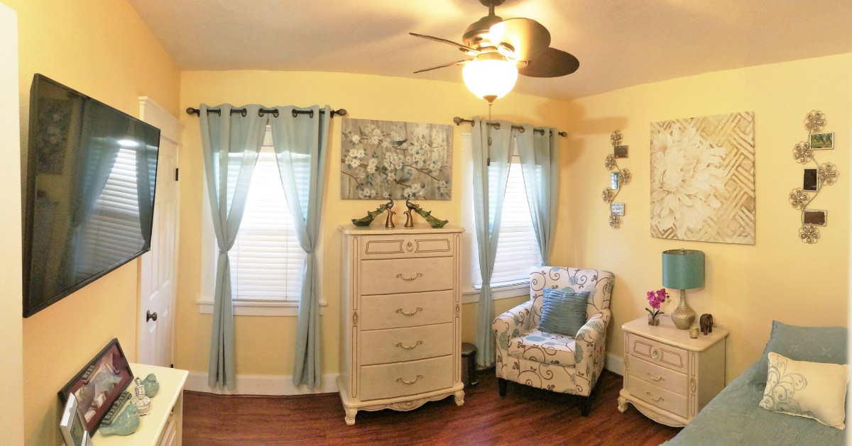 A cozy bedroom with light yellow walls, featuring a white dresser, floral armchair, and blue curtains.