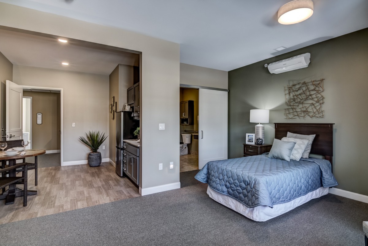 A modern, minimalist bedroom with neutral tones, featuring a cozy bed, a nightstand with a lamp, and an air conditioner on the wall.