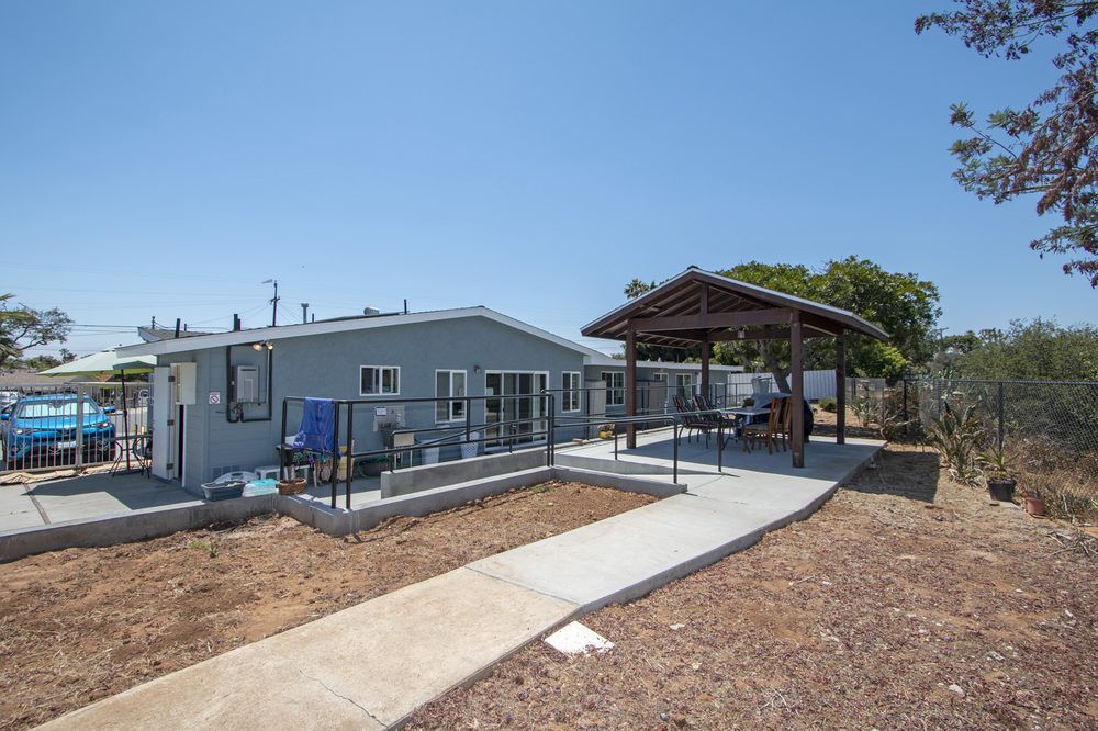 A small house with a paved walkway leading to a covered pavilion in a sunny yard.