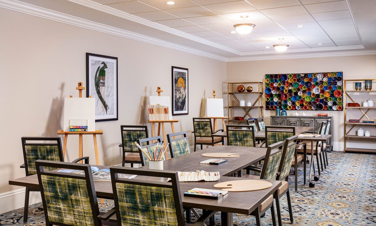 A well-lit art room with tables, chairs, easels, and vibrant paintings on the walls.