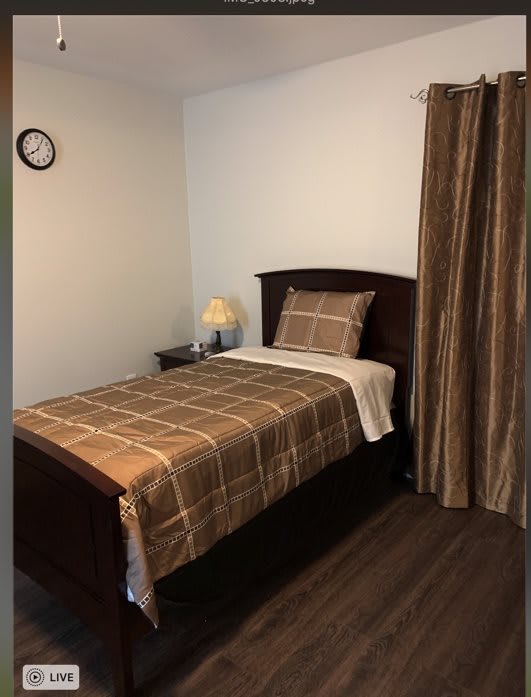 A simple bedroom with a single bed and brown plaid bedding, accompanied by a nightstand, lamp, and wall clock.