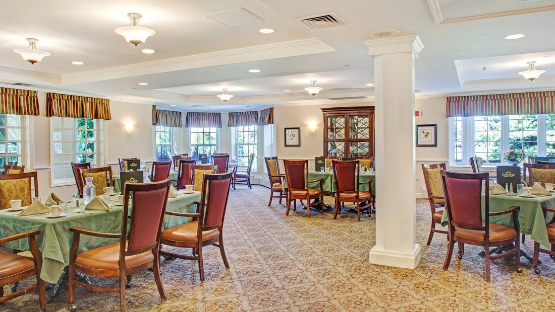 A well-lit dining room with multiple tables and chairs, featuring large windows and a carpeted floor.