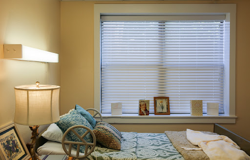 A cozy bedroom featuring a neatly made bed, decorative pillows, and a window adorned with blinds, complemented by personal photographs on display.