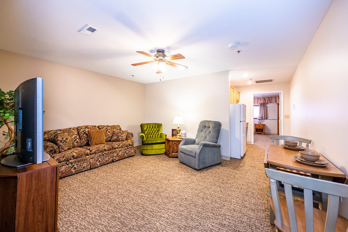 A cozy living room featuring a patterned sofa, two armchairs, a ceiling fan, and carpeted flooring.
