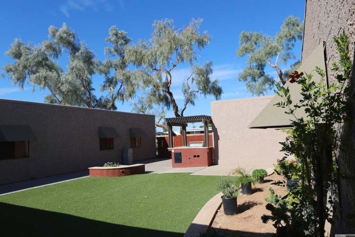 A serene outdoor courtyard featuring artificial grass, brick structures, and lush trees under a clear blue sky.