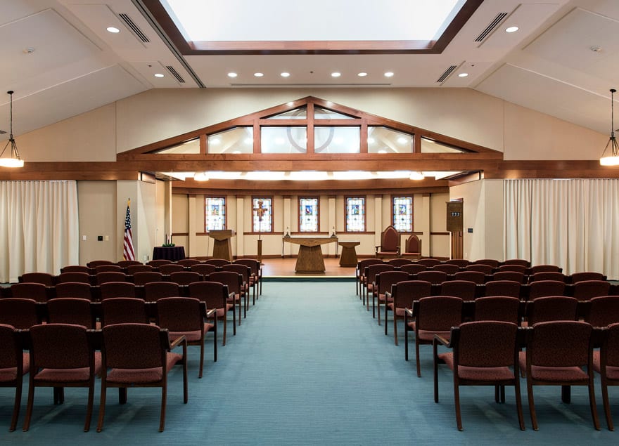 A chapel interior with rows of seats, wooden beams, and large windows.